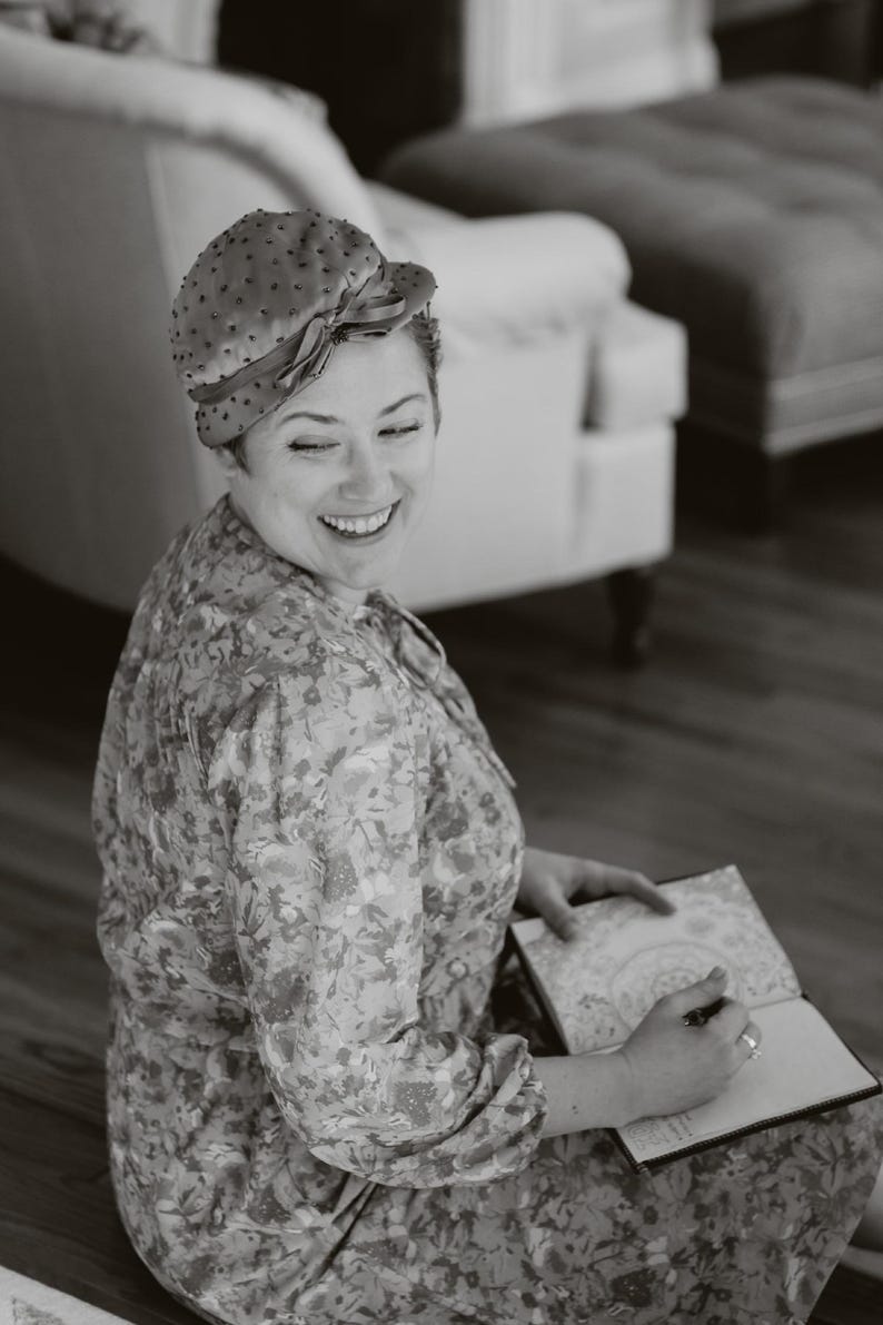 May include: A woman wearing a floral print dress and a dotted hat with a bow, smiling while writing in a notebook. The image is in black and white, with a soft focus, suggesting a vintage or artistic style.