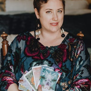 May include: A woman holding a deck of tarot cards. She is wearing a patterned top with floral designs in shades of pink and teal. The cards feature colorful illustrations. She is seated in a wooden chair.