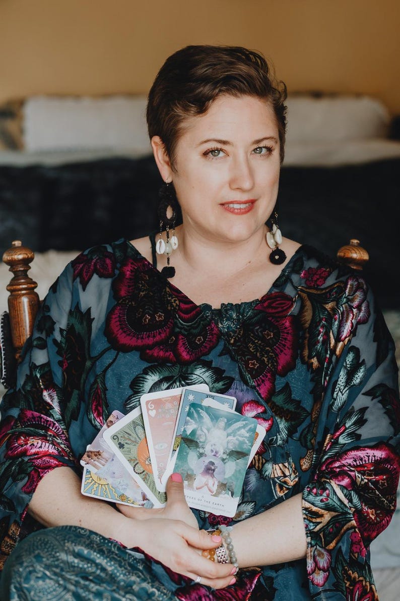 May include: A woman holding a deck of tarot cards. She is wearing a patterned top with floral designs in shades of pink and teal. The cards feature colorful illustrations. She is seated in a wooden chair.