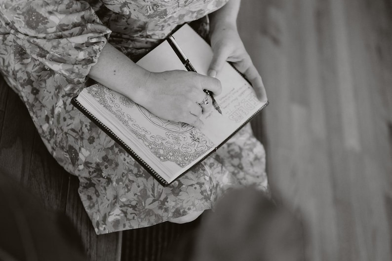 Peut inclure: Une personne &eacute;crit dans un carnet noir avec un stylo. Le carnet est ouvert, r&eacute;v&eacute;lant des dessins d&eacute;taill&eacute;s et des notes manuscrites. La personne porte une robe &agrave; motifs floraux.