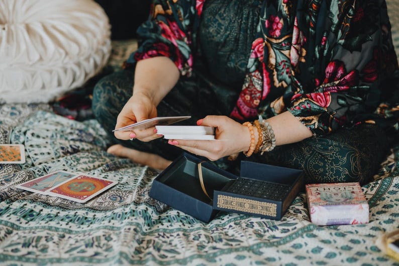 Pode incluir: Uma pessoa segurando cartas de tar&ocirc;, com uma caixa azul e um baralho de cartas vis&iacute;veis. A pessoa est&aacute; vestindo um top com estampa floral e pulseiras. A cena &eacute; ambientada em um cobertor estampado.