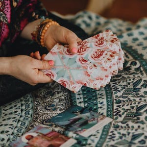 May include: A deck of tarot cards with a floral rose pattern in pink and white is held in the hands. The cards are fanned out, revealing the detailed design. Other cards are visible on a patterned fabric.