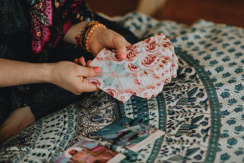 May include: A deck of tarot cards with a floral rose pattern in pink and white is held in the hands. The cards are fanned out, revealing the detailed design. Other cards are visible on a patterned fabric.