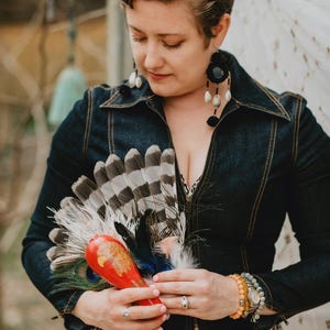 May include: A denim jacket with a collared neckline and a zipper closure. The person is holding a red and orange decorative item with feathers and a peacock feather. The person is wearing large black earrings and a leopard print dress.