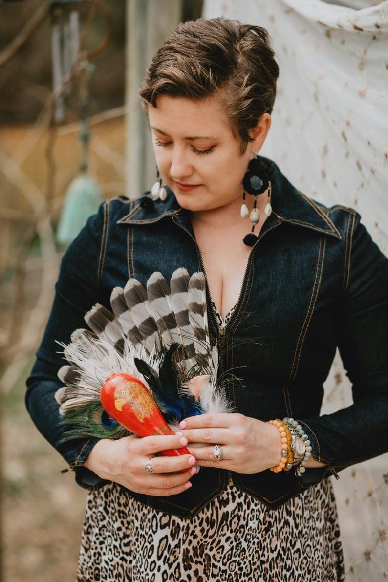 May include: A denim jacket with a collared neckline and a zipper closure. The person is holding a red and orange decorative item with feathers and a peacock feather. The person is wearing large black earrings and a leopard print dress.