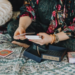 May include: A person is holding a deck of tarot cards, with a blue box and a small box labeled "Light" nearby. The person is wearing a floral patterned top and bracelets. The scene is set on a patterned blanket.