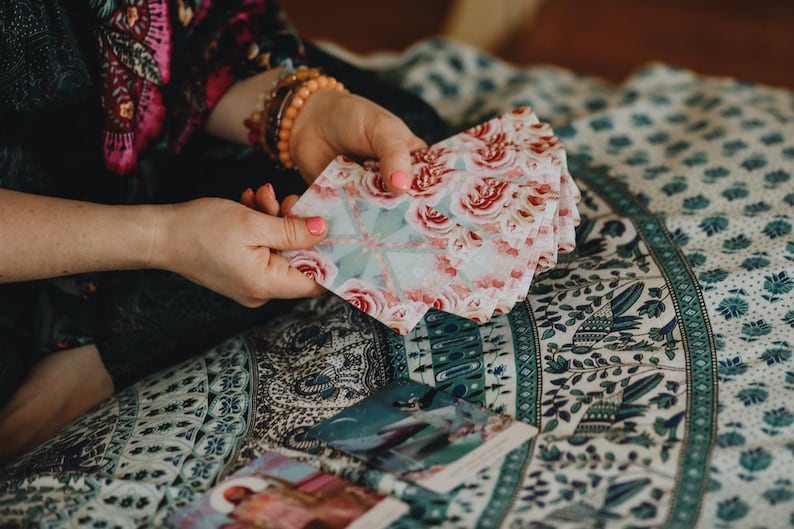 Puede incluir: Un mazo de cartas de tarot con un dise&ntilde;o floral de rosas rosas y blancas se sostiene en las manos. Las cartas est&aacute;n abanicadas, revelando la obra de arte. El fondo presenta un textil estampado con motivos florales turquesa y blancos.