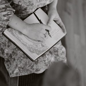 May include: A person writing in a black-bound journal with a pen. The journal is open, revealing detailed line art and handwritten notes. The person is wearing a floral patterned dress.