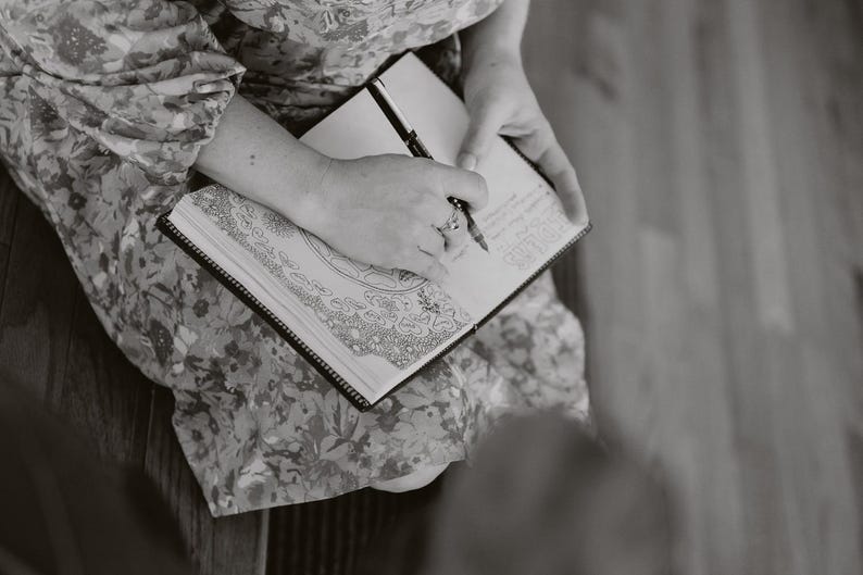 May include: A person writing in a black-bound journal with a pen. The journal is open, revealing detailed line art and handwritten notes. The person is wearing a floral patterned dress.