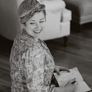 Peut inclure: Une femme souriante, v&ecirc;tue d'une robe &agrave; motifs floraux et d'un chapeau &agrave; pois avec un n&oelig;ud, &eacute;crit dans un carnet. L'image en noir et blanc a une mise au point douce, sugg&eacute;rant un style vintage ou artistique.