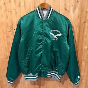 Puede incluir: Una chaqueta universitaria de satén verde con el logotipo de los Philadelphia Eagles bordado en el pecho izquierdo. La chaqueta tiene cierre frontal con botones a presión, mangas largas y puños y dobladillo de punto acanalado con rayas blancas y grises.