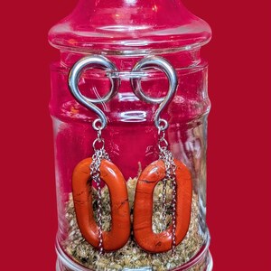May include: A pair of earrings with a silver hook and chain, featuring a reddish-orange, oval-shaped stone. The earrings are displayed inside a clear glass jar with a red background.