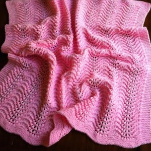 May include: A soft, pink knitted blanket with a wavy pattern and decorative openwork details. The blanket is folded, showing the texture and design. The edges are finished with a scalloped border.