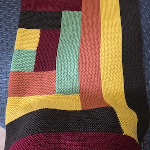 Könnte beinhalten: Eine handgestrickte Decke mit Patchwork-Design. Die Decke zeigt rechteckige Blöcke in verschiedenen Farben, darunter Grüntöne, Gelb, Orange, Burgunderrot und Schwarz. Die Strickstruktur ist deutlich sichtbar und trägt zur gemütlichen Ästhetik bei.