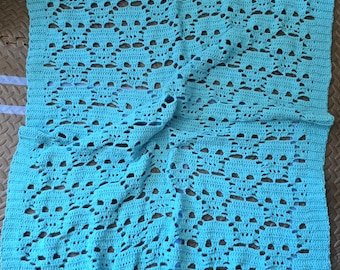 Puede incluir: Una manta turquesa tejida a ganchillo con un patrón de calaveras repetido. La manta está hecha de hilo suave y tiene una apariencia texturizada. El diseño calado crea un textil transpirable y decorativo.