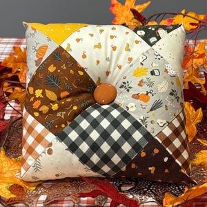 May include: A square patchwork pillow with a button center, featuring autumn-themed fabrics in brown, cream, and black-and-white checkered patterns. The pillow is surrounded by colorful fall leaves on a plaid tablecloth, creating a seasonal display.