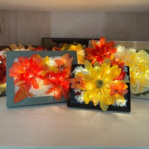 May include: Several framed floral arrangements with LED lights. The flowers are in shades of orange, yellow, and white. The frames are various colors, including brown, black, and teal. The arrangements are displayed on a white surface.