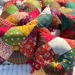 May include: A collection of colorful, handmade patchwork pillows. Each pillow features a unique combination of patterned fabrics in shades of red, green, yellow, and orange, with a button in the center. The pillows are arranged on a white, textured surface.