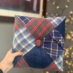 May include: A square decorative pillow constructed from a patchwork of various fabrics, including red and black plaid, blue and white plaid, and solid navy quilted sections. A fabric-covered button sits at the center, and a small metal button is visible.