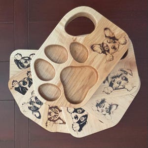 May include: Wooden serving tray with a paw print design and four circular cutouts. The tray has a handle and is decorated with engraved images of various dog breeds. The wood has a natural, light brown color.