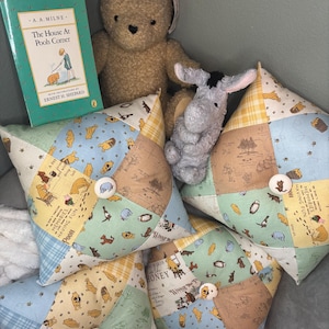May include: Three square patchwork pillows featuring Winnie the Pooh character prints in blue, yellow, green, and tan. Each pillow has a central white button. A book titled The House At Pooh Corner, a tan teddy bear, and a gray donkey plush are present.