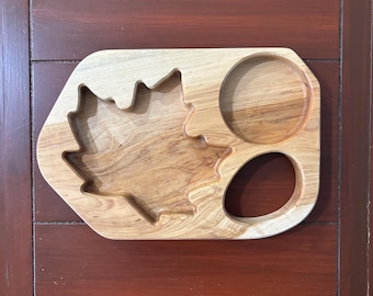 May include: A wooden serving tray with a maple leaf cutout and two circular indentations. The tray is made of light-colored wood with a natural grain pattern. The tray is approximately 10 inches long and 6 inches wide.