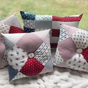 May include: A collection of decorative throw pillows in red, white, and blue patchwork patterns. The pillows feature various designs, including stars, stripes, and floral motifs. Each pillow has a button in the center. The pillows are arranged on a white, textured surface.
