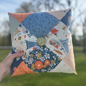 May include: Square patchwork pillow with a button in the center. The pillow features various fabric patterns, including floral, camping-themed, and geometric designs in shades of blue, orange, and cream. The text "Stay Wild" is visible on one of the fabric patches.