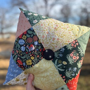 May include: A handmade, square pillow with a patchwork design. The pillow features various floral patterns in shades of pink, blue, green, and yellow. A large, dark blue button is centered on the pillow. The pillow is held in a person's hand.