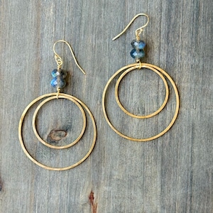 May include: A pair of gold-toned dangle earrings featuring two interlocking circular hoops of different sizes. Each earring is suspended from a gold-toned hook and accented with two small, faceted, blue-grey beads positioned above the hoops.