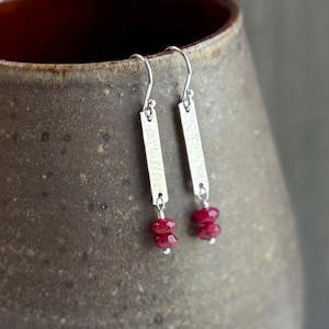 May include: A pair of silver earrings with a hammered texture. Each earring features a rectangular bar with a small, dangling cluster of two red gemstones. The earrings are suspended from a curved hook.