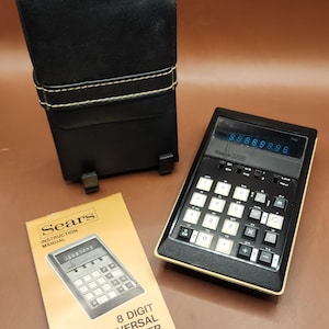 May include: A black 8-digit universal converter calculator with a digital display, a numeric keypad, and function keys. It includes a black protective carrying case with a snap closure and a tan instruction manual labelled Sears 8 Digit Universal Converter.