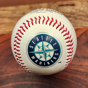 May include: A white Major League Baseball with red stitching featuring the Seattle Mariners logo in the center. The logo is a dark blue circle with a teal and silver compass rose design and the text Seattle Mariners printed in white lettering.