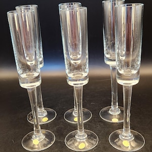 May include: Six clear glass champagne flutes with a unique double-walled design. The flutes have a tall, slender shape with a long stem and a round base. The glasses are arranged on a dark surface, showcasing their elegant form.
