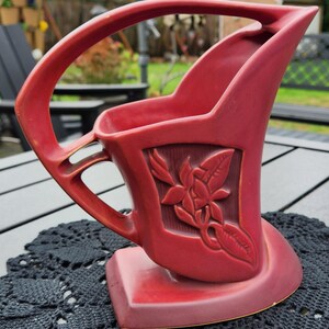 May include: A maroon-colored ceramic vase with a handle and a floral relief design. The vase has a unique shape with a wide base and a curved handle. The floral design is in a rectangular panel on the front of the vase.