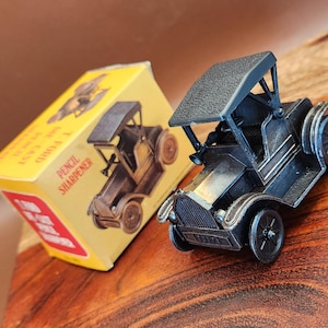 May include: A dark metallic gray die-cast metal pencil sharpener shaped like a vintage T-Ford automobile. It is accompanied by a yellow cardboard box featuring a graphic illustration of the car and the text T-FORD DIE-CAST PENCIL SHARPENER.