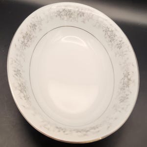 May include: White ceramic bowl with a floral pattern around the rim. The bowl has a gold trim and a silver band. The floral design is in shades of gray. The bowl is empty and sits on a black surface.