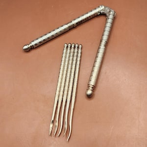 May include: Silver-toned nutcracker and six nut picks arranged on a brown surface. The nutcracker is open, showing its hinged design. The picks are slender with curved tips, and all pieces have a textured pattern.