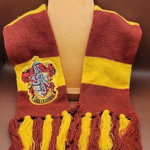 Peut inclure: Écharpe tricotée bordeaux et or, ornée du blason de Gryffondor. L'écharpe a un corps bordeaux avec des rayures et des glands dorés. Le blason de Gryffondor est brodé sur l'écharpe. L'écharpe est présentée sur une forme en bois.
