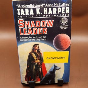 May include: A paperback book titled "Shadow Leader" by Tara K. Harper. The cover features a woman in a cloak, a wolf, and a planet. The book is labeled "Autographed" with the SF logo.