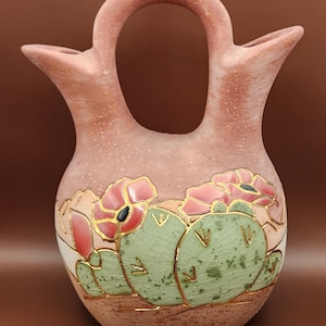May include: A handcrafted, reddish-brown pottery vase with a unique handle and two spouts. The vase features a painted design of green cacti and pink flowers with gold accents. The vase has a textured surface and a speckled appearance.