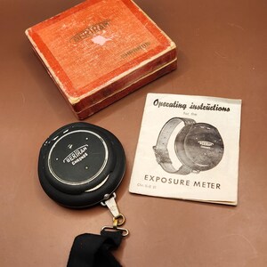 May include: Vintage black Bertram Chronos exposure meter with a black strap, a red box, and an instruction manual. The meter has the text "BERIRAM CHRONOS" on its face. The instruction manual reads "Operating instructions for the EXPOSURE METER".