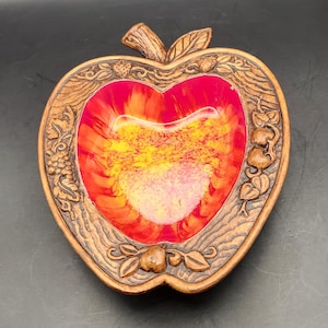 May include: A decorative apple-shaped dish with a carved wooden frame. The interior features a vibrant red, orange, and yellow design. The frame is adorned with carved leaves, grapes, and other fruit details.