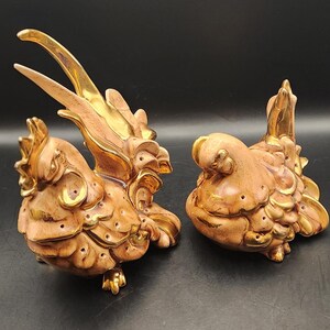 Könnte beinhalten: Zwei Keramik-Hahnfiguren. Die Hähne sind pfirsichfarben mit goldenen Akzenten auf Flügeln, Kämmen und Füßen. Ein Hahn steht aufrecht, während der andere in Ruheposition ist. Die Figuren haben eine detaillierte, strukturierte Oberfläche.