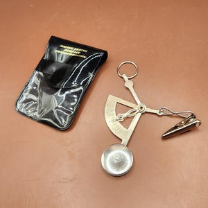 May include: A vintage metal instrument with a circular base, a curved scale, and a clip, next to a black vinyl case. The case has the text "BOEING VERTOL SURFACE TRANSPORTATION" in gold lettering.