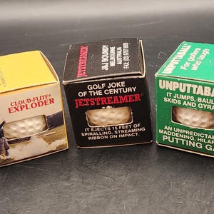 May include: Three novelty golf ball boxes: yellow "Cloud-Flite Exploder", black "Jetstreamer", and green "Unputtaball". Each box contains a golf ball and humorous text. The boxes are designed as gags.