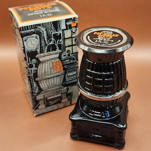 May include: A black glass bottle shaped like a pot-belly stove with a silver-toned rim and a circular label on top reading Avon Pot-Belly Stove Bravo After Shave 5 fl. oz. It is accompanied by a matching cardboard box with a black and white illustration.