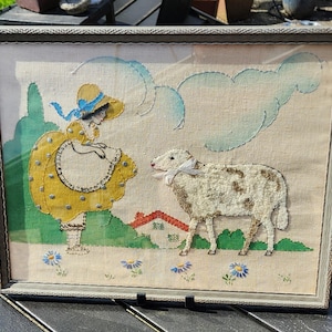 May include: Framed vintage textile art featuring a girl in a yellow dress and a sheep. The artwork includes blue accents, a red roof, and a light-colored background. The frame is a neutral color.