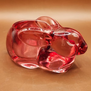May include: A clear, red glass bunny-shaped container. The container has a rounded body with a bunny head and ears. The glass is smooth and reflects light, creating highlights and shadows. The container is likely a decorative item or a small holder.