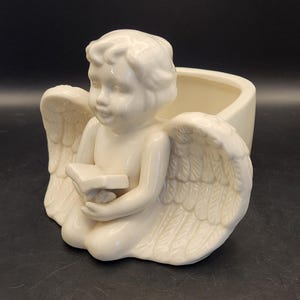 May include: Cream-colored ceramic angel planter. The angel is kneeling, holding a star-shaped object, with large feathered wings. The planter has a rectangular opening for plants or other decorative items. The angel is a decorative item.